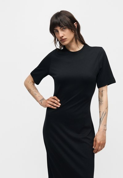 Γυναίκα με σκούρα μαλλιά και τατουάζ στα χέρια, που φοράει εφαρμοστό μαύρο φόρεμα με κοντά μανίκια, ποζάρει με το ένα χέρι στη μέση της μπροστά από απλό φόντο.