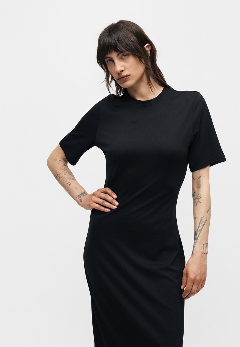 Frau mit dunklen Haaren und Tätowierungen an den Armen, die ein eng anliegendes schwarzes Kleid mit kurzen Ärmeln trägt und mit einer Hand in der Hüfte vor einem einfarbigen Hintergrund posiert.