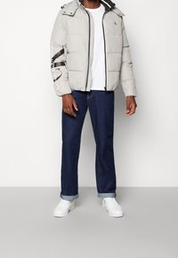 Giacca puffer grigio chiaro con zip frontale, cappuccio regolabile e logo sulla manica sinistra, abbinata a jeans in denim blu e sneakers bianche.