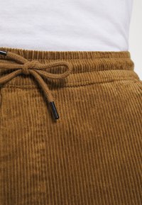 Bruine corduroy broek met een tailleband en een strik van het koord, gedragen met een wit shirt erin gestopt.