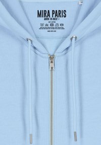 Haut à capuche zippé bleu clair en 100 % coton. Présente une fermeture éclair à l'avant, des cordons de serrage et une étiquette avec des instructions d'entretien à l'intérieur.