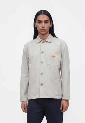 Homme aux longs cheveux noirs portant une veste beige clair boutonnée avec trois poches avant et une petite étiquette logo sur la poche poitrine.