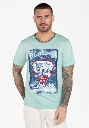 Man met kort haar en tatoeages draagt een lichtgroen T-shirt met de opdruk "Raceway 94 Bikers" in blauw, rood en wit, staand tegen een witte achtergrond.