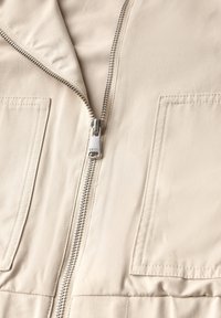 Beige zipjacka med slät tygtextur, som har två stora fackfickor och silverdetaljer på dragkedjan.