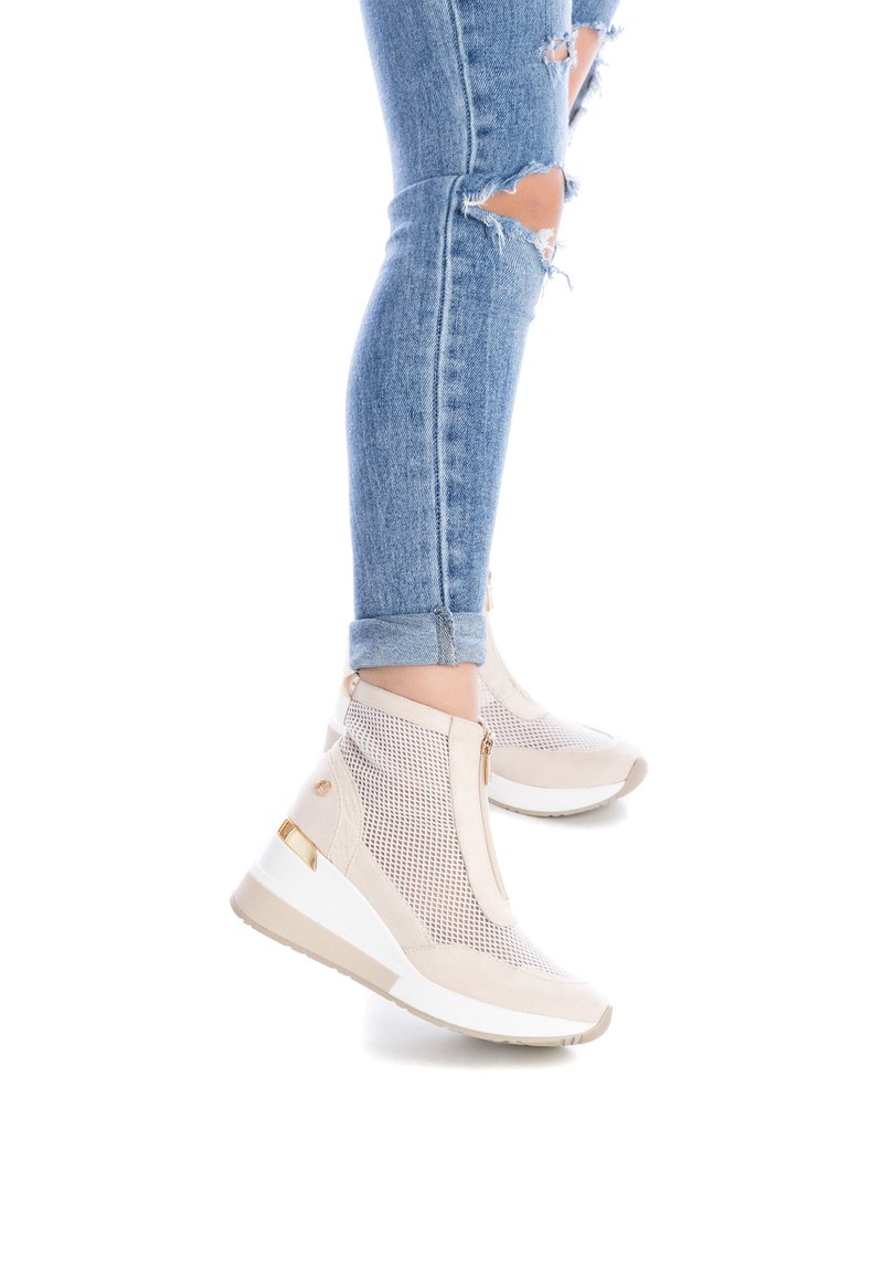 Zapatillas cuña beige con paneles de malla, acentos dorados y cremalleras laterales. La suela es blanca con un agarre texturizado. Los jeans tienen rodillas desgastadas.
