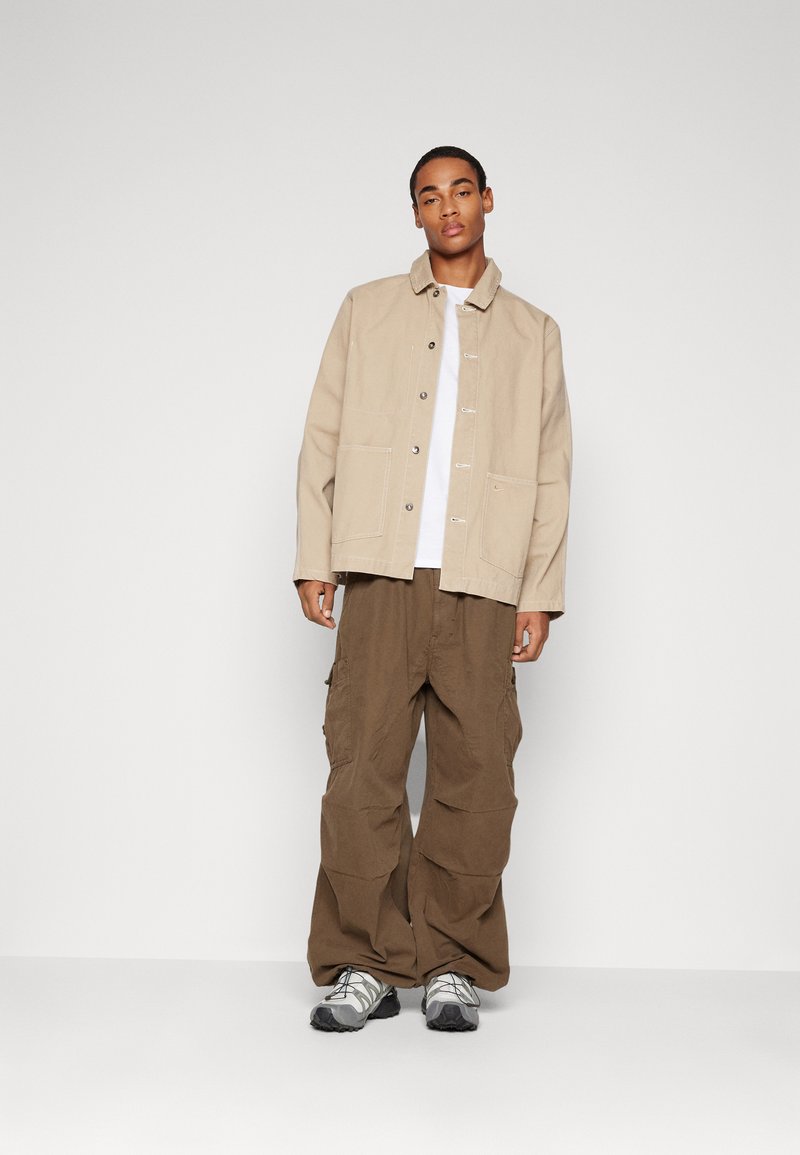 Chaqueta de algodón beige con cierre de botones en la parte delantera y dos bolsillos frontales, combinada con pantalones cargo marrones de corte holgado y zapatillas grises.