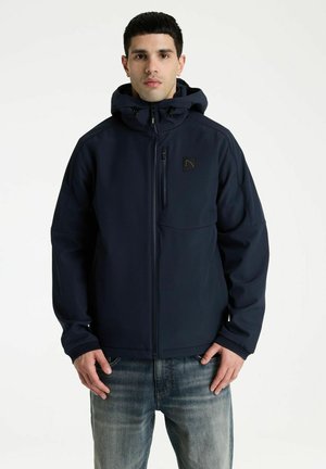Man wearing a dark blue hooded zip-up jacket and blue jeans standing against a plain white background.
