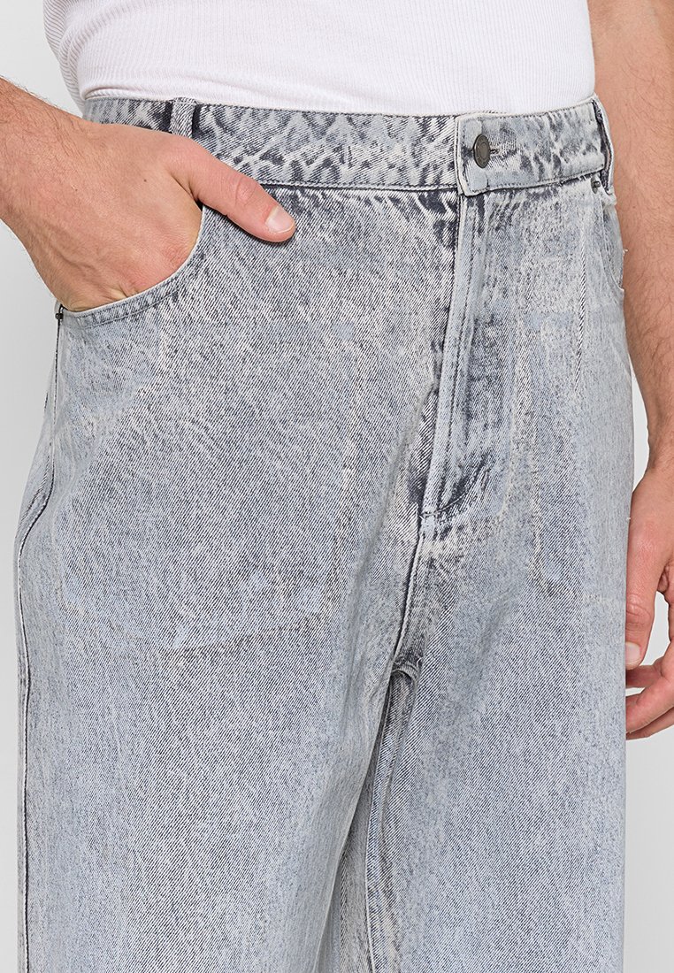 Man in een lichte grijze acid-wash jeans met zijn linkerhand in zijn zak en een wit geribbeld mouwloos shirt in zijn broek gestopt.