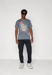 Harmaa t-paita, jossa on graafinen kuva leijonasta ja auringosta, yhdistetty mustiin housuihin, päällä aurinkolasit ja harmaat lenkkarit.