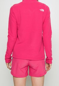 Fleecetröja i klar rosa med hög krage, långa ärmar och logga på ryggen; ihop med matchande rosa shorts. Slät textur.