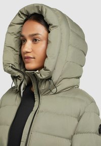 Vrouw in een lichtgroene gewatteerde winterjas met een capuchon, voorzien van een zwarte rits en koordjes, kijkt lichtjes naar links.