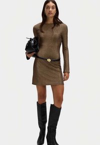 Robe longue en bronze scintillant à manches longues avec une silhouette ajustée, associée à une ceinture noire et des bottes noires jusqu'au genou, tenant un sac noir.