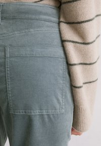 Groene stoffen broek met een achterzak met gestikte details. Een beige gestreepte trui is gedeeltelijk zichtbaar aan de linkerkant.