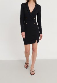 Schwarzes langärmliges Wickelkleid mit tiefem V-Ausschnitt, goldenen Knöpfen und einem Schlitz vorne. Kombiniert mit schwarzen, rückenfreien High Heels.