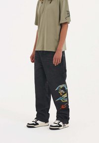 Svarta denim cargo-byxor med drakbroderi i blått och rött, kombinerade med en lös olivgrön t-shirt i bomull och svarta och vita sneakers.