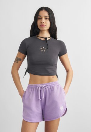 Noor naine, kellel on pikad tumedad juuksed, kannab tumedat halli lühikest t-särki lilledetailiga ja heleroosasid paelaga lühikesi pükse, käed taskutes.