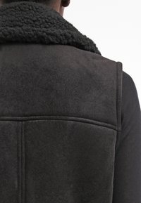 Persona che indossa una giacca shearling nera con dettagli della cucitura sulla schiena e un colletto strutturato, di spalle su uno sfondo bianco.