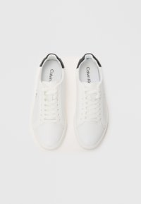 White sneakers with a smooth texture, featuring black accents at the heel, flat laces, and a minimalistic design. Brand name inside.