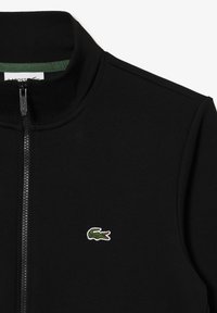Veste noire à fermeture éclair avec un col haut et un petit logo crocodile vert sur le côté gauche de la poitrine.