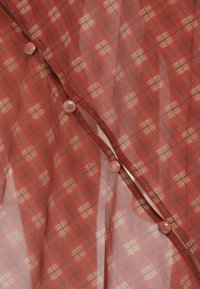 Läpinäkyvä punainen kangas, jossa on ruudullinen kuosi kevyillä vivahteilla. Mukana koristepainikkeet reunassa.