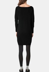 Donna che indossa un vestito nero a maniche lunghe, lungo fino al ginocchio, collant neri, calze grigie e scarpe nere, in piedi di spalle contro uno sfondo semplice.