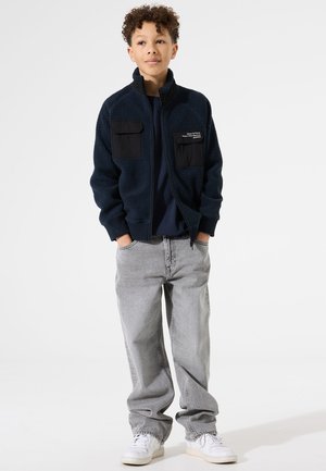 Marineblaues Fleecesakko mit hohem Kragen, zwei vorderen Taschen und schwarzen Akzenten, kombiniert mit grauen Denim-Hosen und weißen Sneakers.
