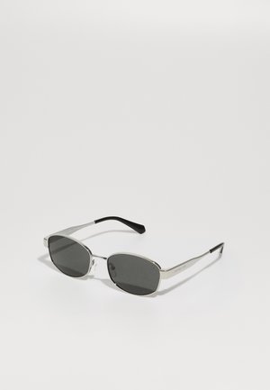 Óculos de sol com lentes cinzas, armação metálica prateada fina, detalhes transparentes e pontas pretas nas hastes. Apresenta um formato oval.