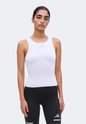 Femme aux cheveux plaqués en arrière portant un haut de sport blanc sans manches et des leggings noirs avec des logos Adidas sur fond blanc.