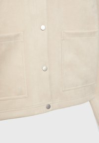 Beżowa zamszowa kurtka z dwoma przednimi kieszeniami, zatrzaskami i gładką fakturą. Detale obejmują srebrne zapięcia i czysty, prosty design.