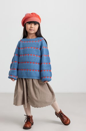 Blauwe gebreide trui met rode harten, beige rok met een gegolfde zoom, roze gebreide maillot en bruine veterschoenen, gecombineerd met een rood gestreepte baret.