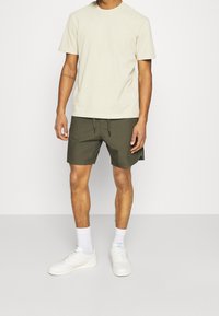 Homme portant un t-shirt beige à manches courtes, un short à cordon olive, des chaussettes blanches et des sneakers blanches, se tenant contre un fond blanc.