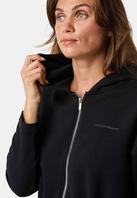 Schwarze Zip-Hoodie aus weichem Stoff, mit einem silbernen Reißverschluss und einem Logo auf der Brust. Die Kapuze ist mit einem passenden Material ausgestattet.