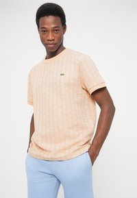 Vyras dėvi persikų spalvos trumpomis rankovėmis marškinį su logotipu ant krūtinės ir šviesiai mėlynas kelnes, stovintis rankas į kišenes padėjęs ant baltos fono.