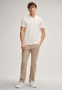 Kurzarm-Polohemd in Off-White mit strukturiertem Muster, kombiniert mit beigen Chinos und weißen Sneakers, stehend vor einem neutralen Hintergrund.