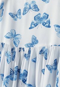 Bílá látka s opakujícím se vzorem motýlů v různých odstínech modré. Texturované volány podél lemu dodávají designu detail.