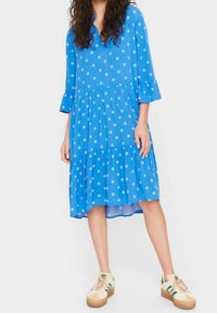 Blå prickig klänning med trekvartsärmar, flerlagersdesign och medellång fåll, kombinerad med avslappnade sneakers. Mjuk tygkänsla.
