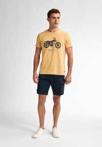 Žluté bavlněné tričko s grafikou motocyklisty, námořnické cargo šortky a bílé tenisky. Jednoduchý design, volný střih a krátké rukávy.