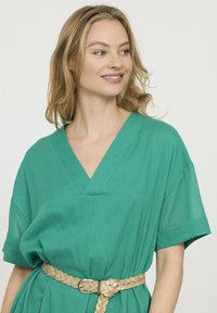 Femme blonde aux cheveux mi-longs portant une robe verte décolletée en V avec des manches retroussées et une ceinture tressée beige, souriant et regardant à droite.