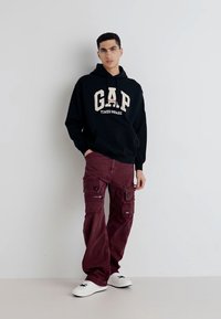 Felpa nera con logo "GAP TIMES SQUARE" in beige, abbinata a pantaloni cargo bordeaux con più tasche e una vestibilità rilassata. Scarpe bianche.