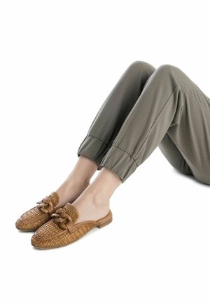 Scarpe slip-on beige con un motivo tessuto e dettaglio a fiocco, abbinate a pantaloni olive a sigaretta, mostrando il piede e la caviglia.