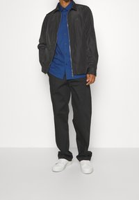 Svart lättvikts zip-up jacka över en blå skjorta med knappar, matchat med svarta byxor och vita sneakers. Jackan har krage och logotypaccent.