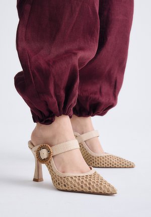 Pies con zapatos de tacón alto beige tejidos con hebillas redondas combinados con pantalones granate sueltos y fruncidos.