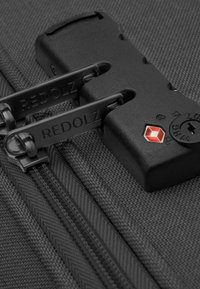 Tirante della zip nero e meccanismo di bloccaggio su tessuto nero strutturato, con logo "REDOLZ" sulle zip e un emblema rosso sul lucchetto.