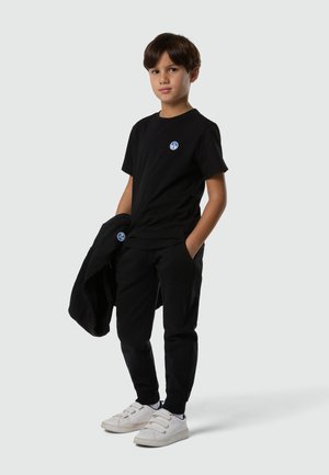 Ragazzo in piedi con una mano in tasca, indossa una maglietta nera e pantaloni, tiene una giacca nera e indossa scarpe da ginnastica bianche su uno sfondo semplice.