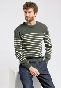 Maglione verde con strisce orizzontali bianche, colletto rotondo e bottoni sulle spalle, abbinato a jeans in denim blu scuro. Morbida texture lavorata a maglia.