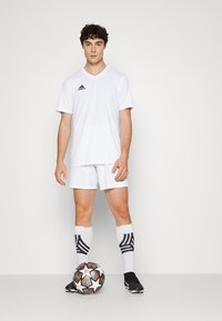 Witte Adidas sporttrui en -shorts. Zwart logo op het shirt. Zwarte voetbalschoenen en kniehoogte witte sokken met zwarte strepen. Voetbalschoen bij de voeten.