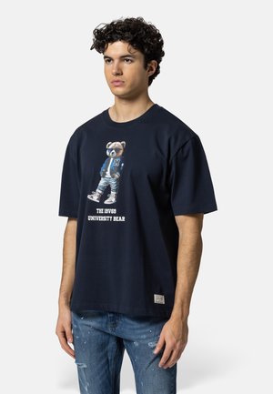 Jonge man draagt een navy T-shirt met een teddybeer in zonnebril en varsityjack, gecombineerd met beschadigde blauwe jeans.