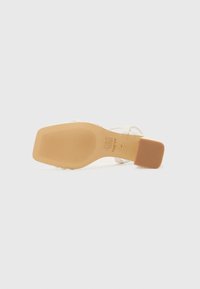 Sandale à talon transparent avec une semelle beige, arborant un design texturé. La partie supérieure est composée de brides transparentes, préservant une esthétique minimaliste.