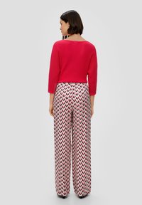 Roter gerippter Pullover mit Dreiviertelärmeln, kombiniert mit weit geschnittenen gemusterten Hosen, die pinke und schwarze Pfeilmuster aufweisen.