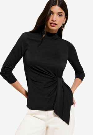 Nő fekete, hosszú ujjú, magas nyakú és oldalán csomós részletű felsőt visel, fehér nadrággal, egyik kezét a csípőjén tartva áll.